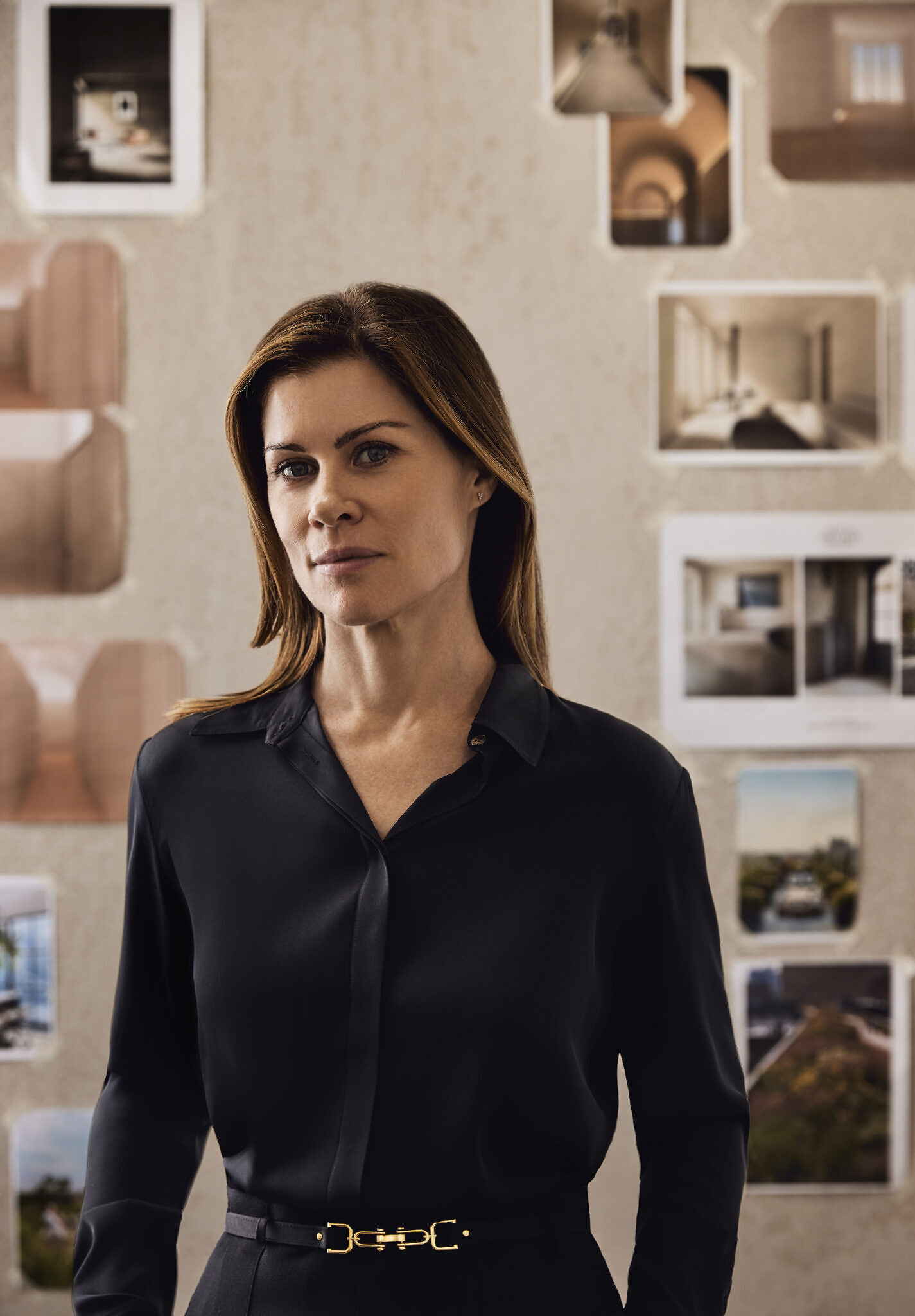 Portrait of Olivia Williams, wearing a black button down shirt with dress pants and a gold and black belt. She has brown hair with blonde highlights. There is a wall behind her of a mood board.