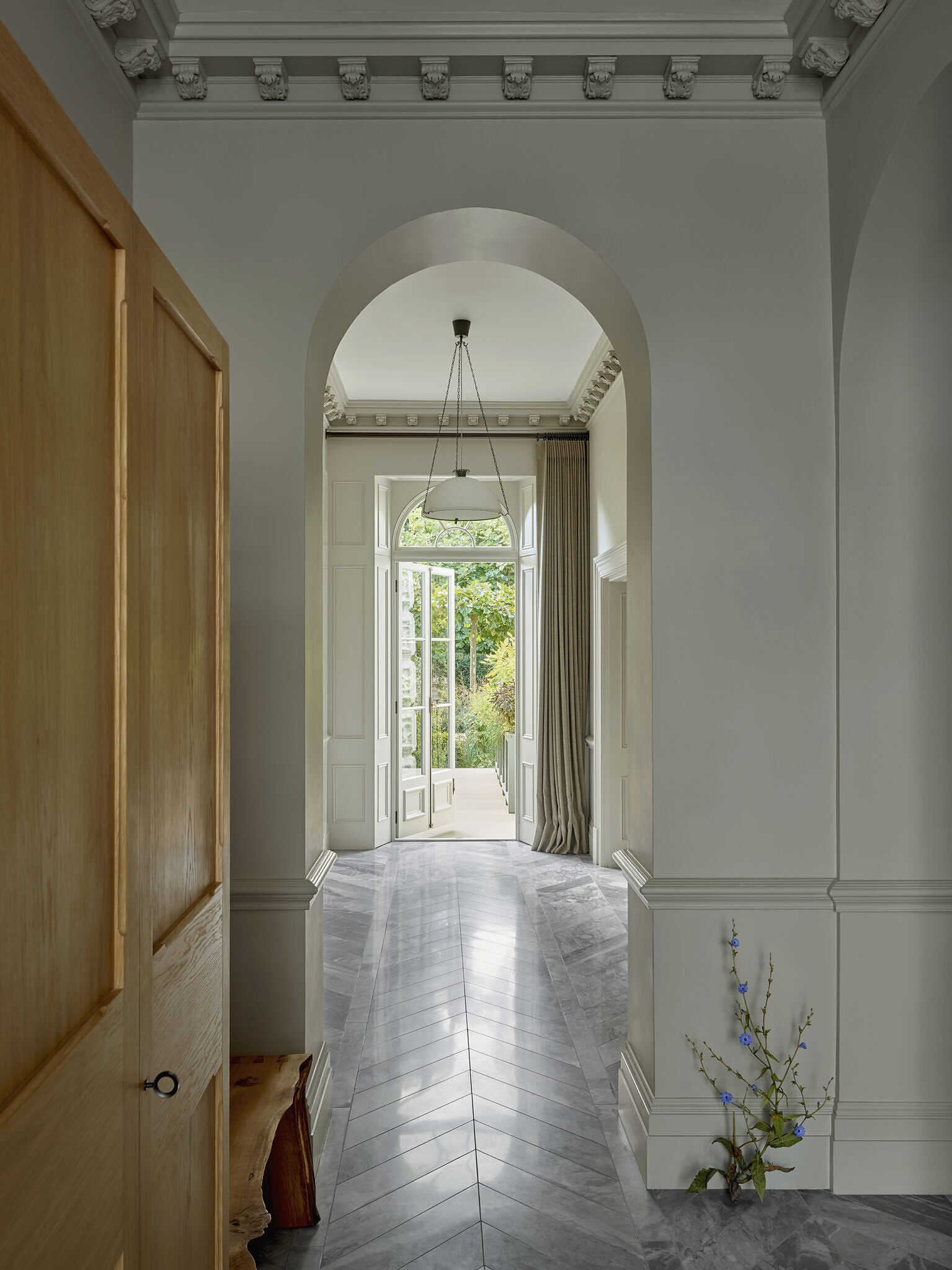 Hallway in the home with a wooden cabinet to the left side, and some flowers on the floor. The door on the opposite side of the image leads to a walkway outside.