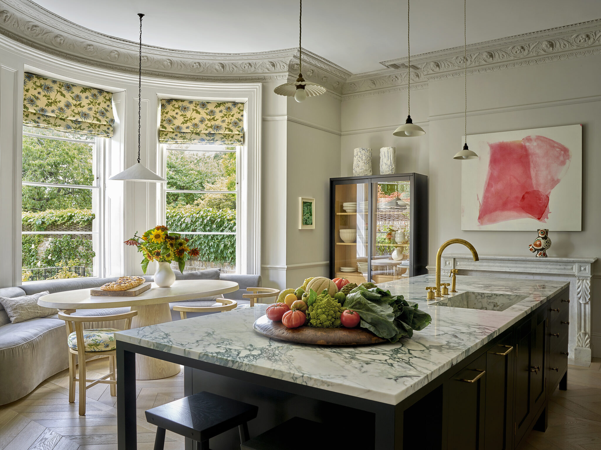 Kitchen image with marble countertops on the kitchen island and gold detailing. There is an oval table behind, with a curved sofa and chairs with flower print.