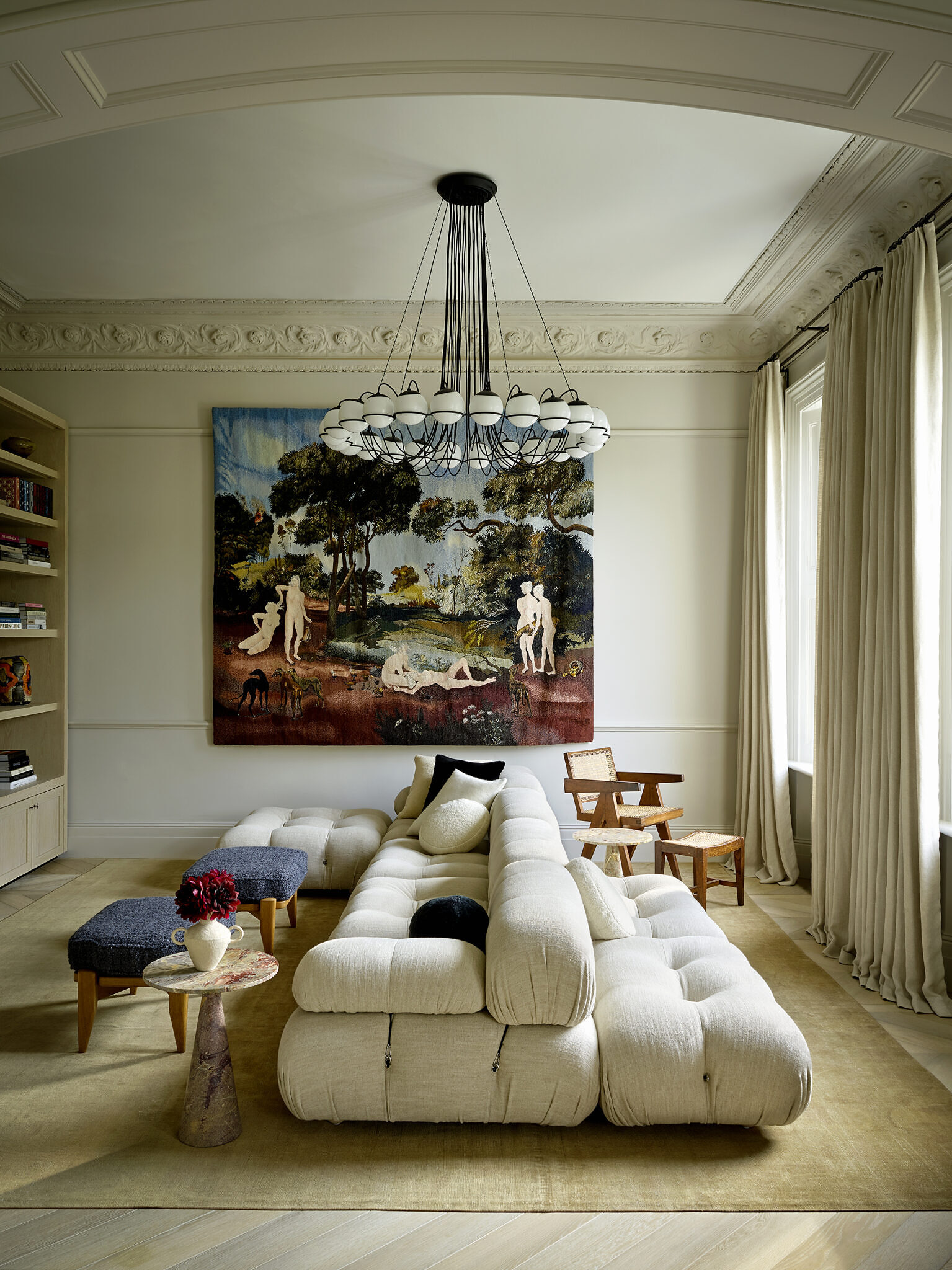 Area of a living room with a large sofa, chairs, and small marble tables. There is a large chandelier which is black with white circular bulbs. There is a large colorful painting on the wall.