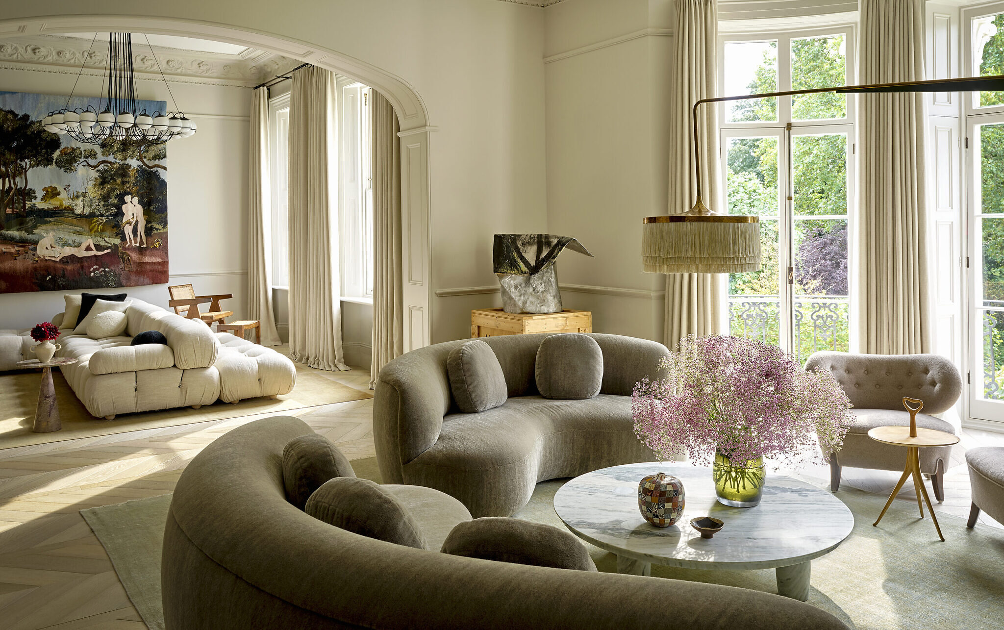 Image of a living room with rounded couches, a marble circular table with flowers in a vase. There is a large lamp overhead. The windows are white and tall, with trees outside.