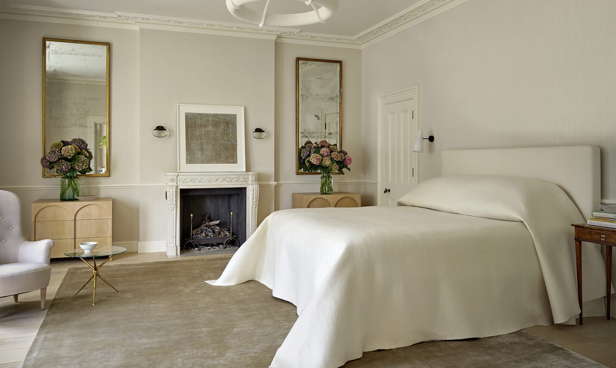 Image of a bedroom which has white bedding, white fireplace, and white ceiling lamp. The drawer units are good with circular detailing.