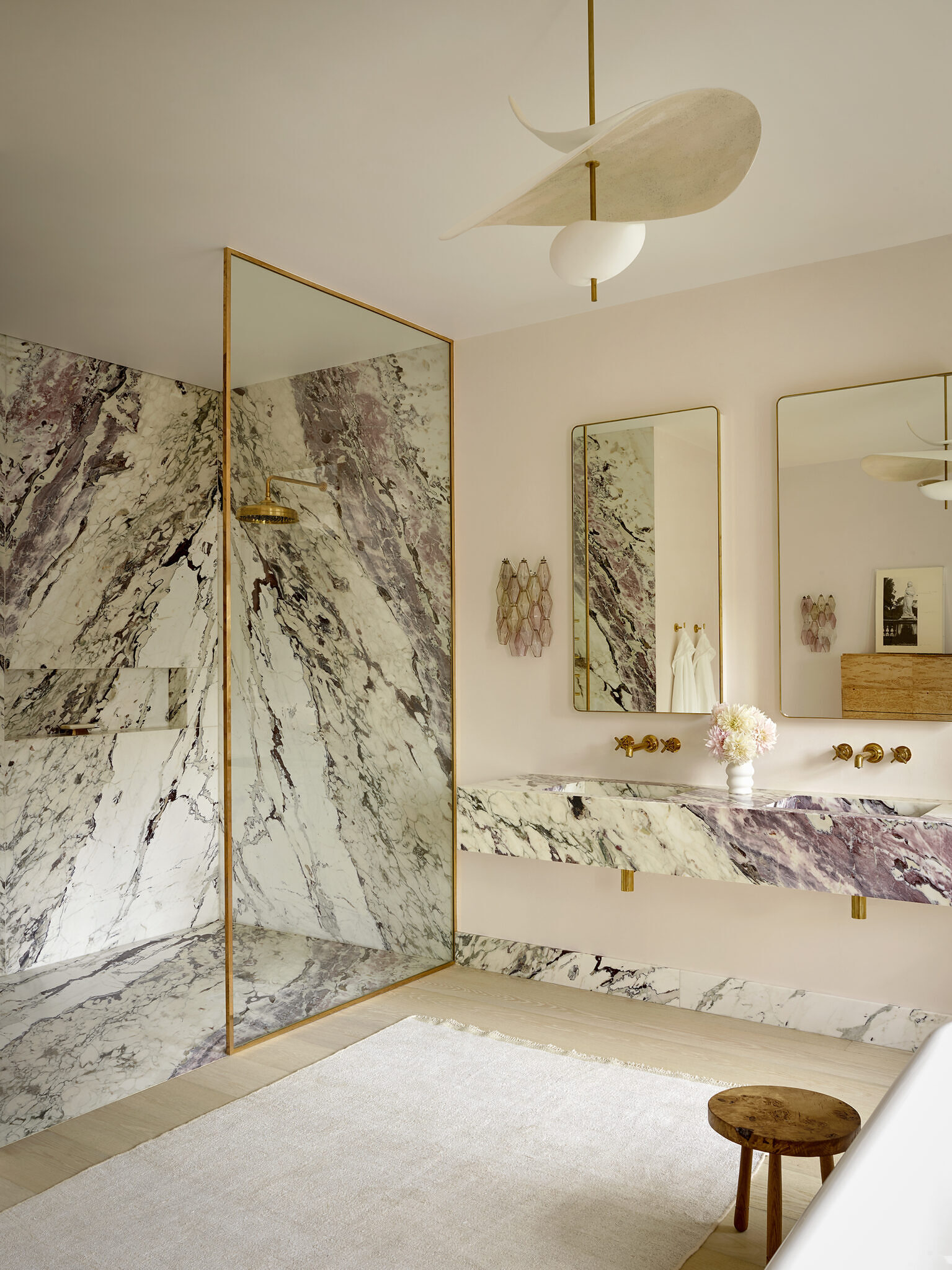 Image of the bathroom, with a marble and gold shower, two tall mirrors and two sinks. There is a light colored rug and a small wooden chair in the center of the bathroom.