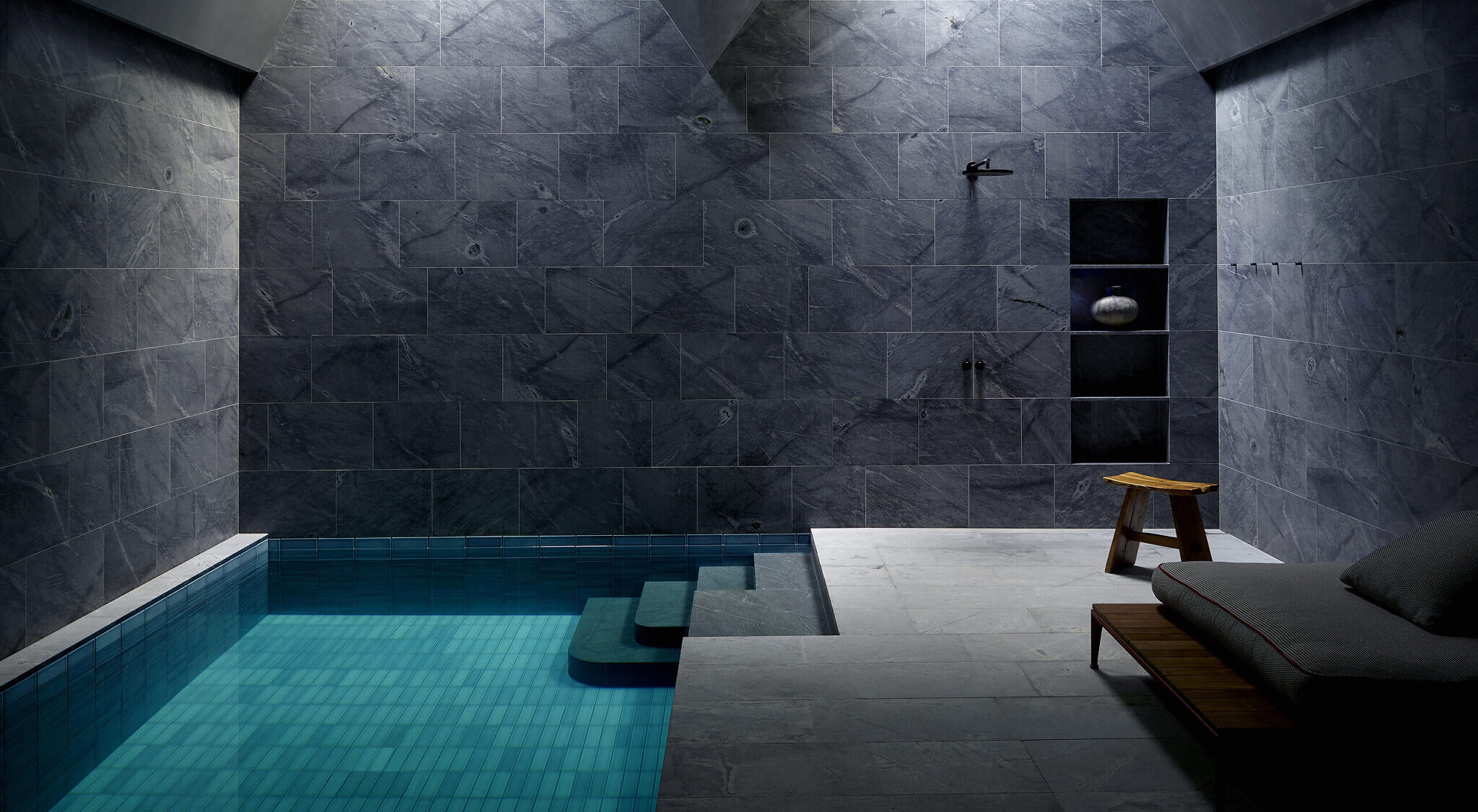 Image of the indoor pool and shower - the walls are grey stone. There is a small wooden chair, as well as a lounge chair on the right side of the image.