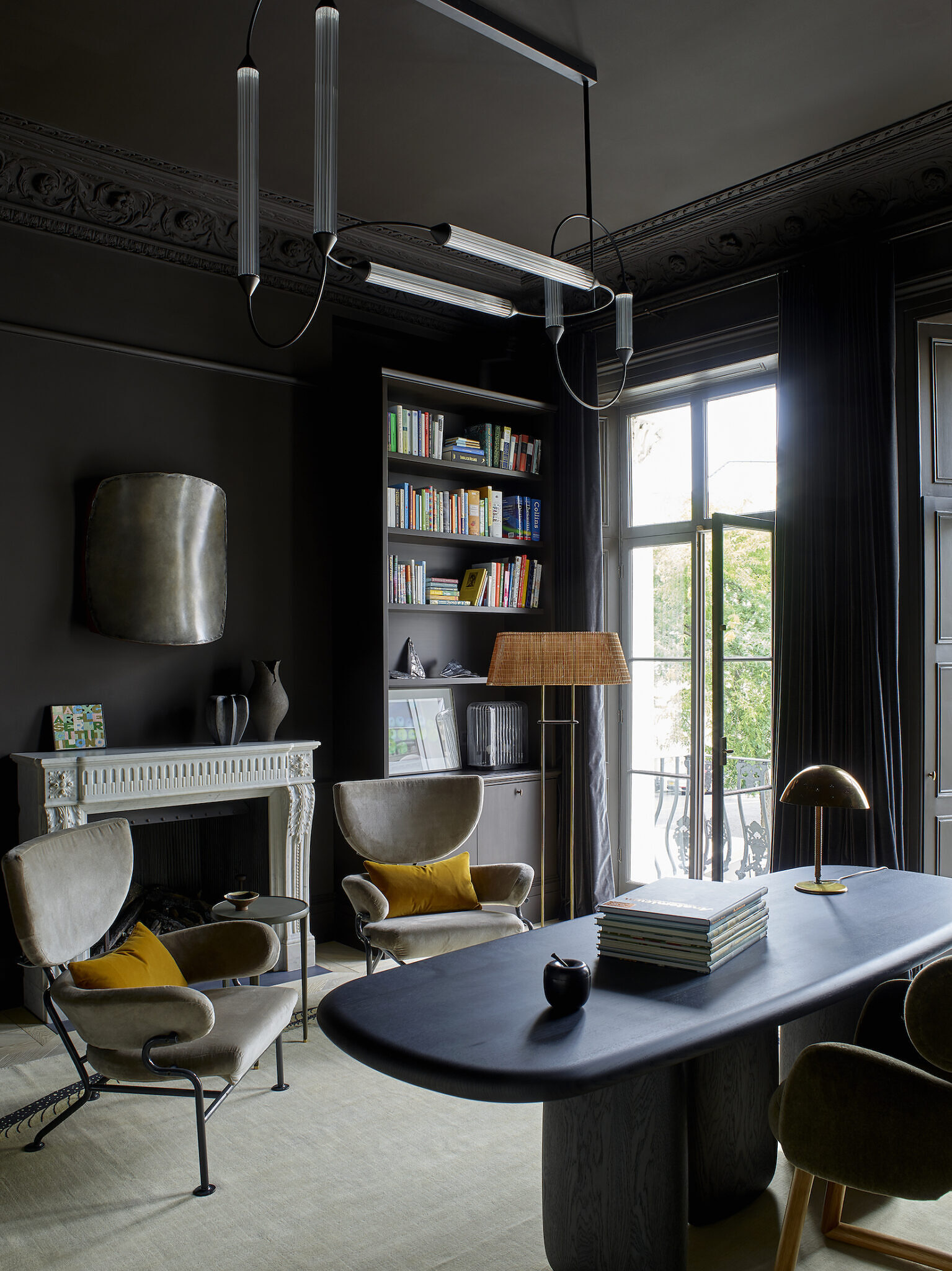 Image of the office space, which has a very large bookshelf, two chairs, and a desk. There are open windows which lead to an outdoor space.