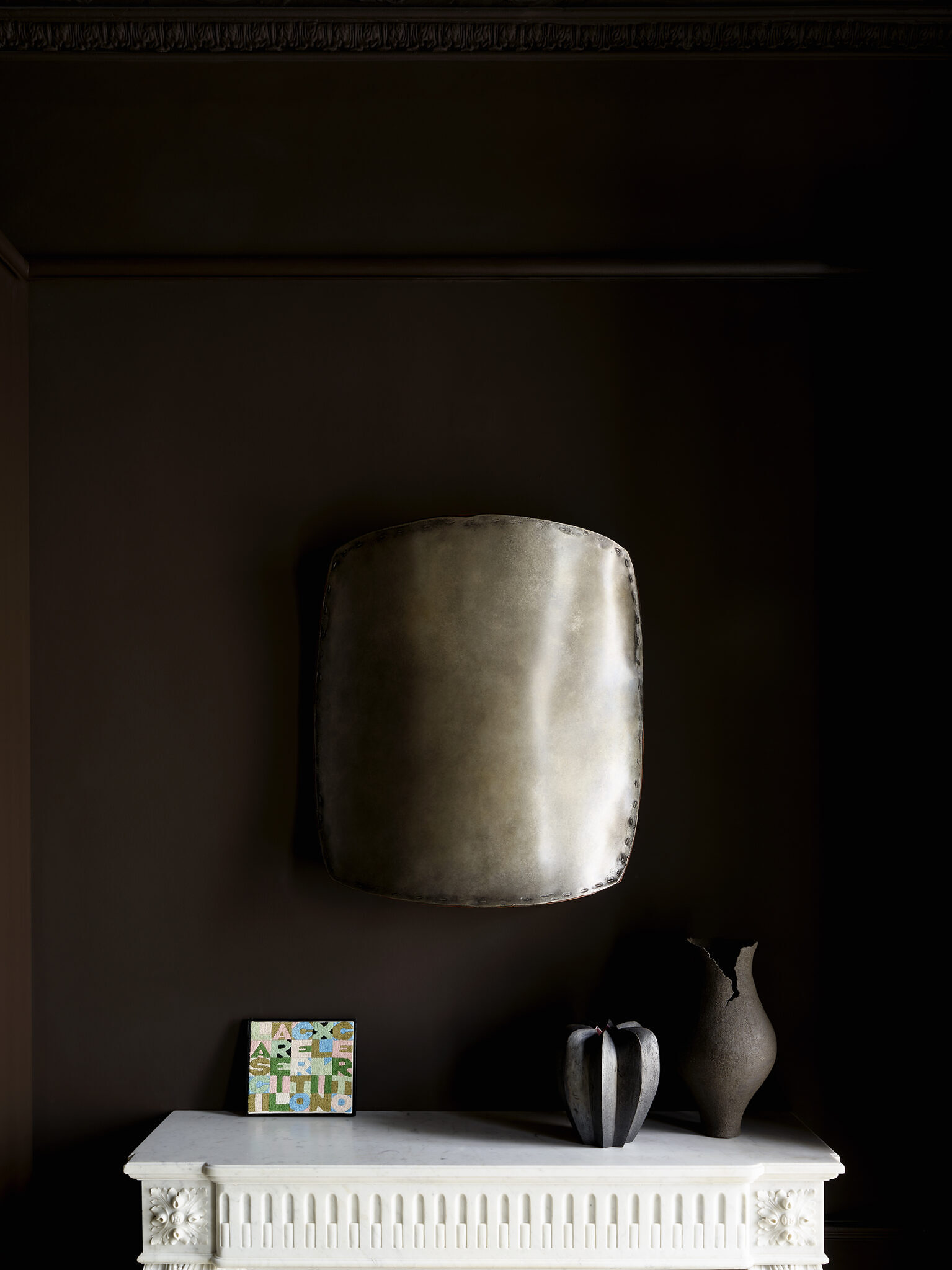 Close up shot of metal shield-like artwork hanging above the fireplace, with a vase and smaller artwork sitting atop the fireplace.