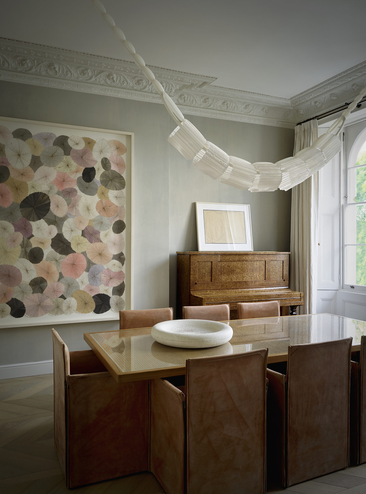 Dining room seating area, with wooden table and seating. White circular ceramin tray is on top of the table. There is a piano off to the side and a large, colorful square piece of artwork on the wall.